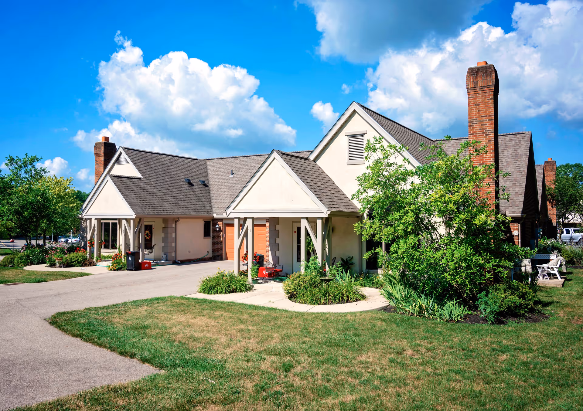 Exterior view of a single-story building with a pitched roof, brick chimneys, and a covered entrance surrounded by greenery and a well-maintained lawn under a partly cloudy blue sky.