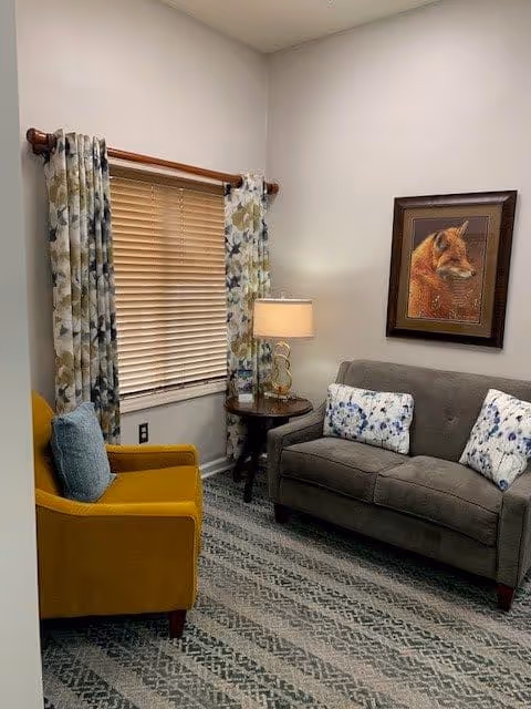 A cozy sitting area with a gray loveseat adorned with two floral pillows, a mustard yellow armchair with a blue pillow, a round wooden side table with a lamp, floral curtains on a window with closed blinds, and a framed picture of a fox on the wall.