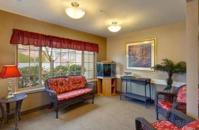 A cozy sitting area with wicker furniture featuring red floral cushions, a small side table with a red lamp, a fish tank on a wooden stand, a framed painting of a forest scene on the wall, and a potted plant. Large windows with red valances let in natural light and show greenery outside.