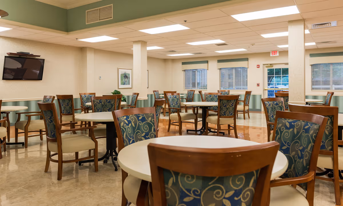 A well-lit dining room with multiple round tables and wooden chairs featuring patterned upholstery. The room has beige walls with green wainscoting, several windows with blinds, a wall-mounted television, and an exit door. The floor is a mix of polished tiles and linoleum.