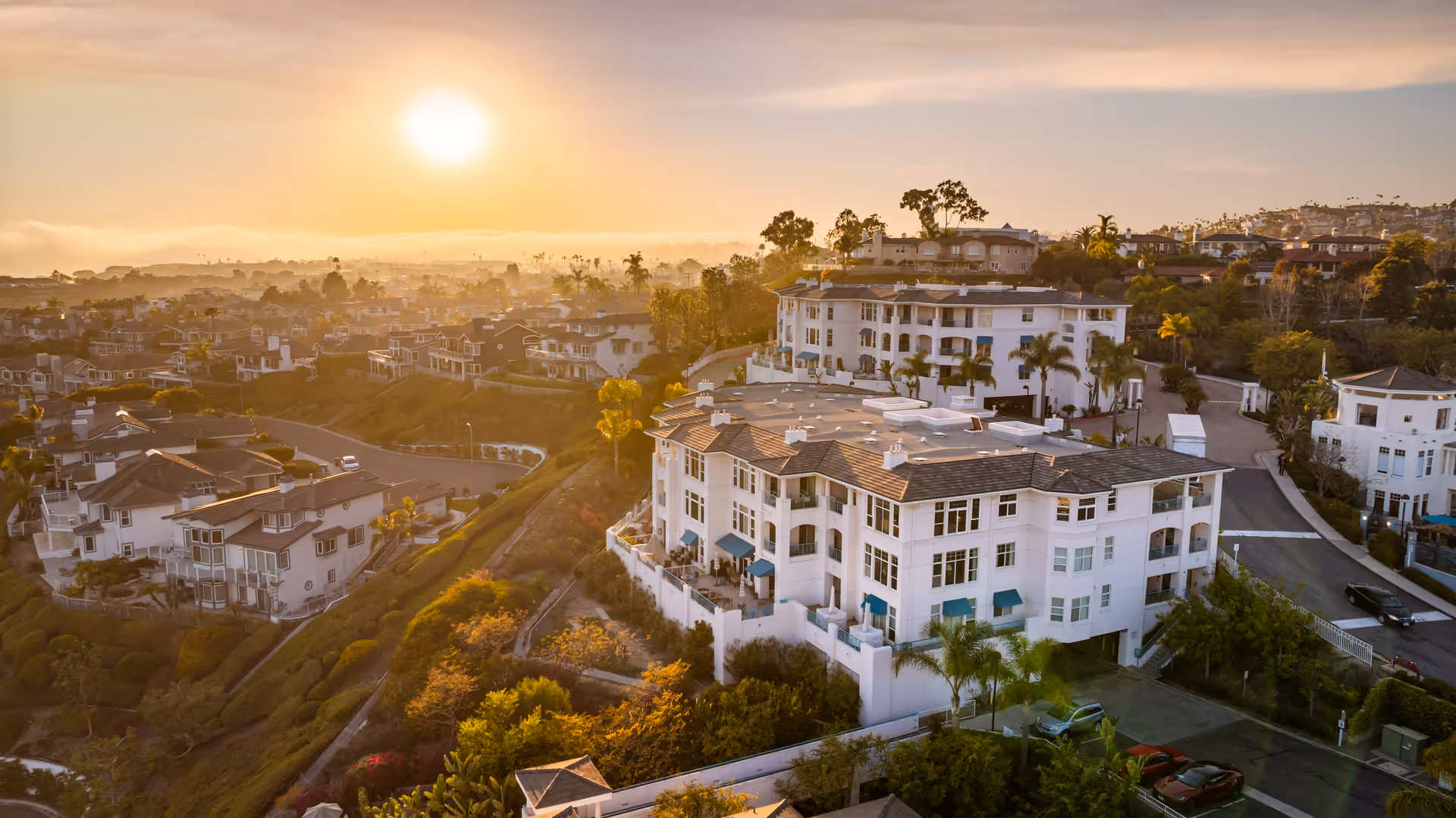 Aerial view of a residential community at sunset with multiple white buildings surrounded by greenery and roads, with the sun low in the sky casting a warm golden light.