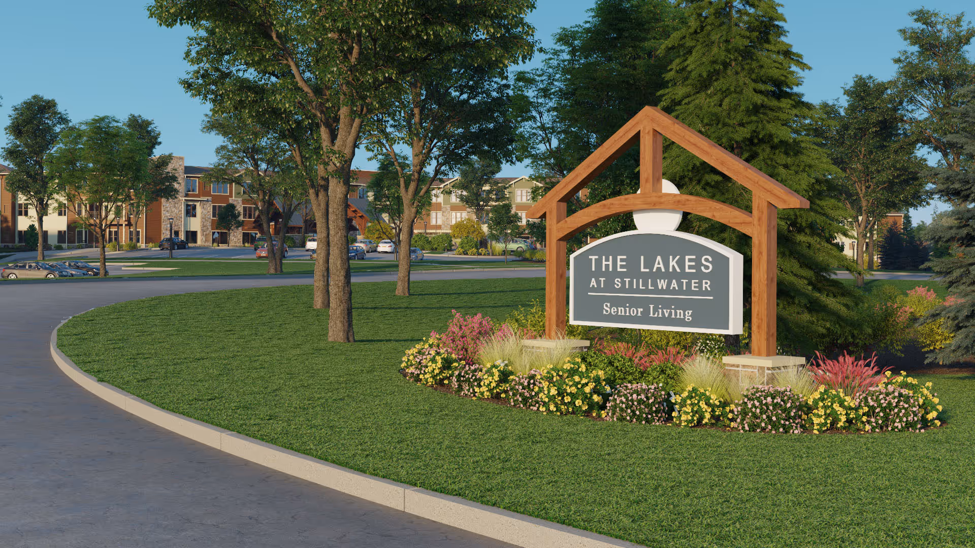 Entrance sign for The Lakes at Stillwater Senior Living facility surrounded by green grass, colorful flowers, and trees with the senior living buildings visible in the background under a clear sky.