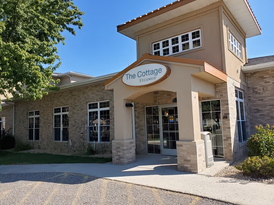 Exterior front entrance of a building named The Cottage Ecumen, featuring beige brick walls, large windows, a covered entryway, and a tree on the left side under a clear blue sky.