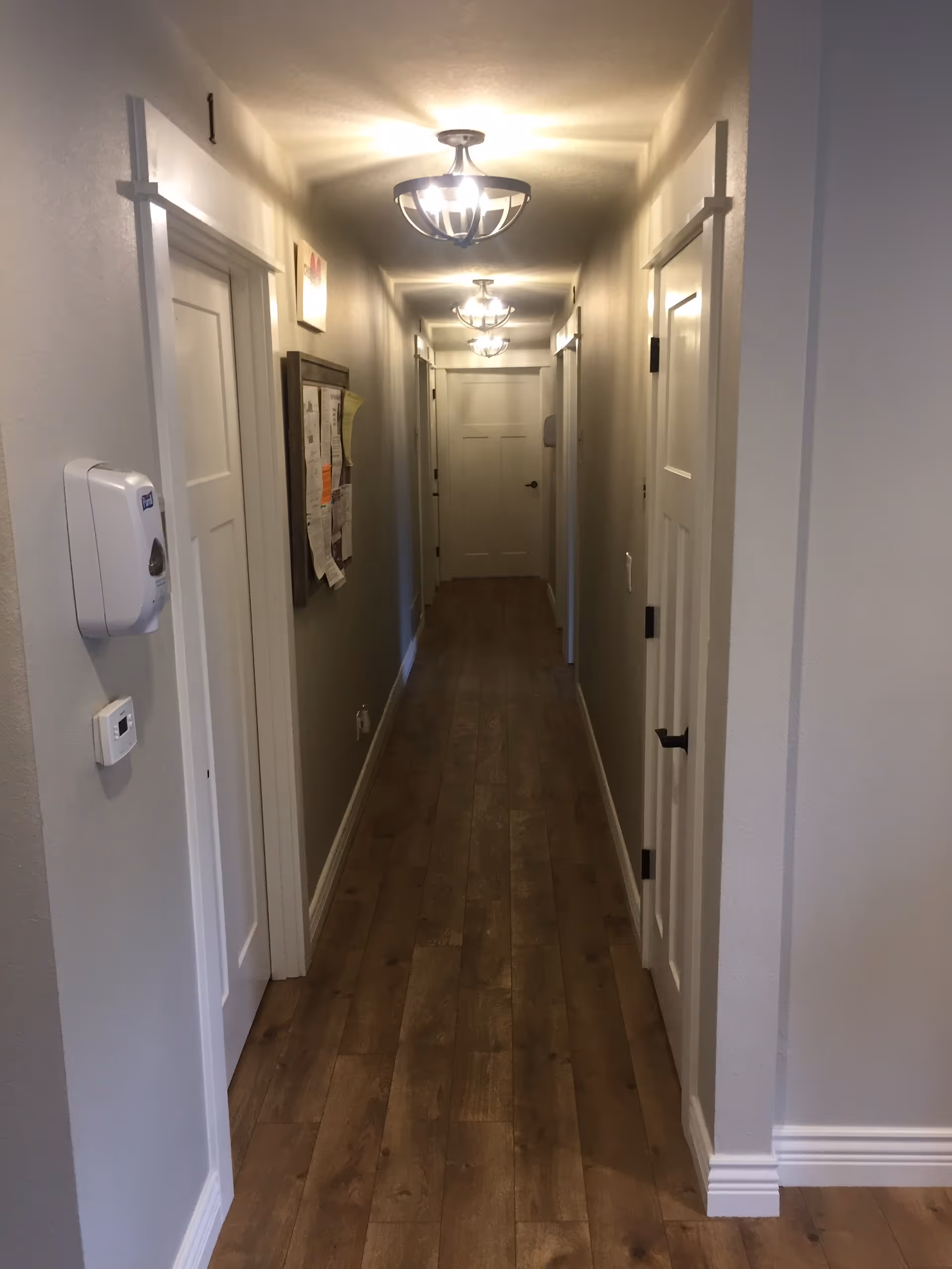 Long, well-lit interior hallway with wood flooring, multiple doors on both sides, ceiling lights and a bulletin board on the wall.