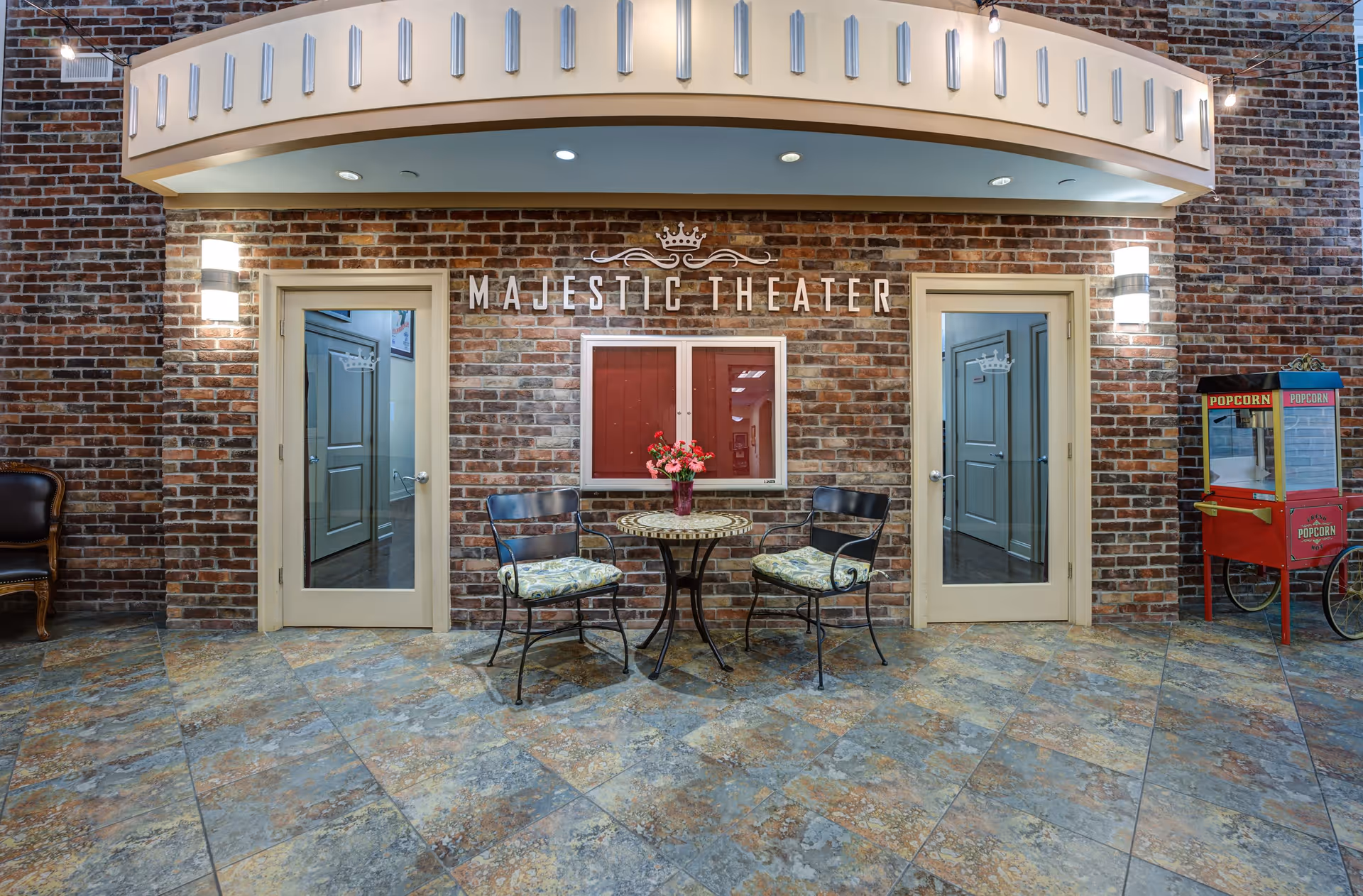 Interior view of a theater entrance area with a brick wall background. Two glass doors flank a central window display with the words 'Majestic Theater' above. In front of the wall, there is a small round table with a flower vase and two cushioned metal chairs. To the right, there is a red popcorn machine on wheels. The floor is tiled with a stone pattern.