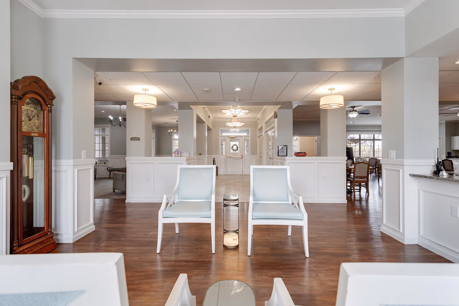A bright and spacious common area in an assisted living facility featuring two light blue cushioned chairs with white frames facing each other, a wooden grandfather clock on the left, and a hallway leading to a door with a wreath. The room has wooden floors, white walls with wainscoting, and ceiling lights.