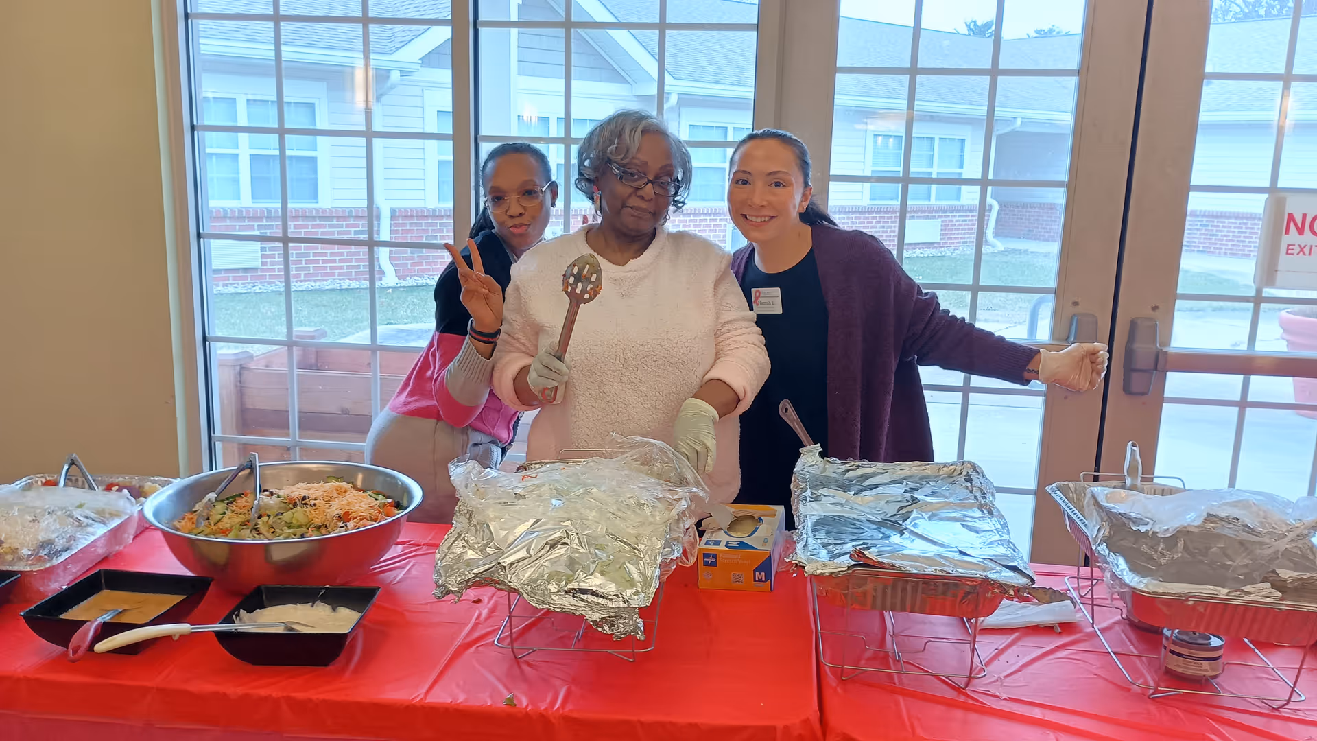 Three women standing behind a table covered with a red tablecloth, serving food from trays covered with aluminum foil. One woman in the center is holding a serving spoon, the woman on the left is making a peace sign, and the woman on the right is smiling. Behind them is a large window and glass door showing the exterior of a building.
