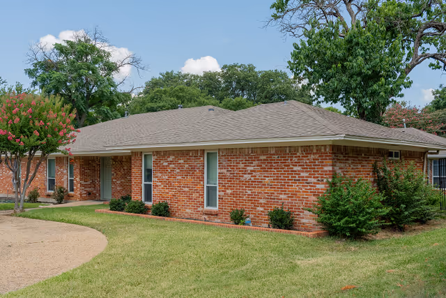 Single-story red brick residential building with a low-pitched roof, landscaped lawn, and trees under a blue sky.