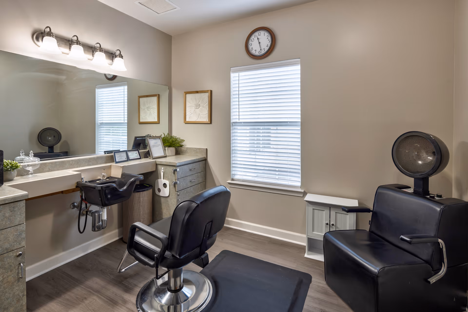 Small salon room with two black styling chairs, a shampoo sink, hood dryer, large mirror and a window.