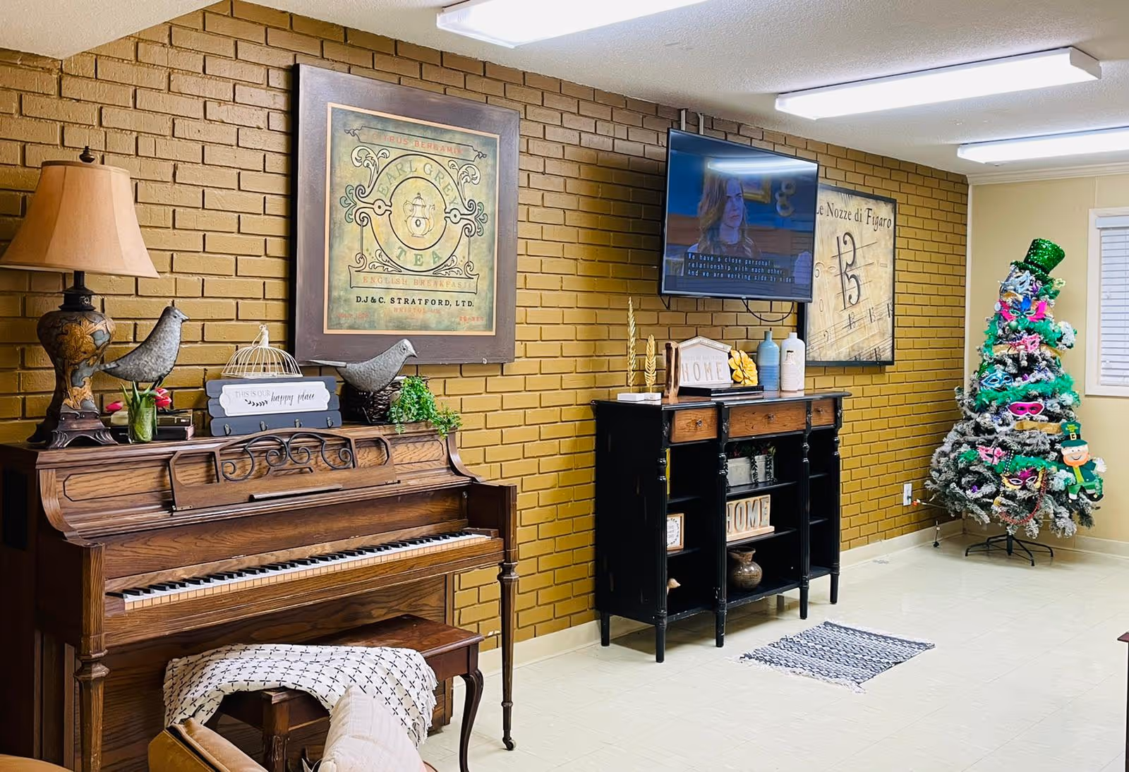 A cozy interior room with a wooden piano adorned with decorative birds, a lamp, and a small sign. Above the piano is a framed vintage-style Earl Grey tea poster. To the right, a black cabinet with shelves holds decorative items and a small 'HOME' sign. A flat-screen TV is mounted on a mustard yellow brick wall, displaying a woman speaking. Further right, a decorated Christmas tree with colorful ornaments and a green top hat stands near a window with white blinds. The floor is tiled and there is a small rug in front of the cabinet.