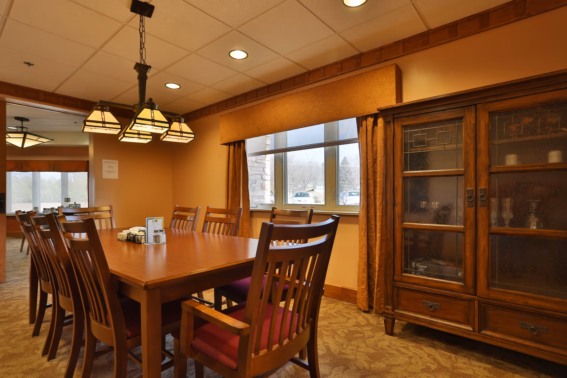 A dining room with a large wooden table surrounded by wooden chairs with red cushions. Above the table hangs a chandelier with multiple square lamps. There is a window with orange curtains and a wooden cabinet with glass doors displaying glassware. The room has warm lighting and beige walls.