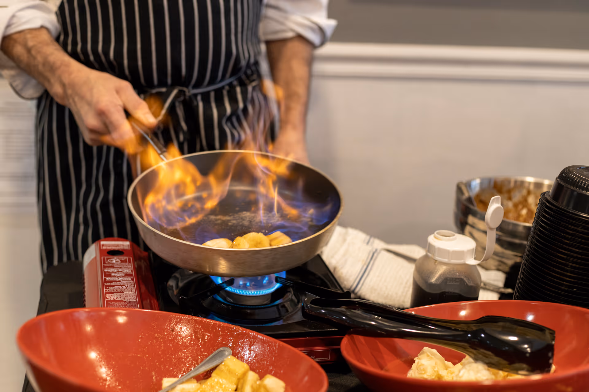 A cook in a striped apron flambés food in a skillet over a portable burner with red serving bowls in the foreground.