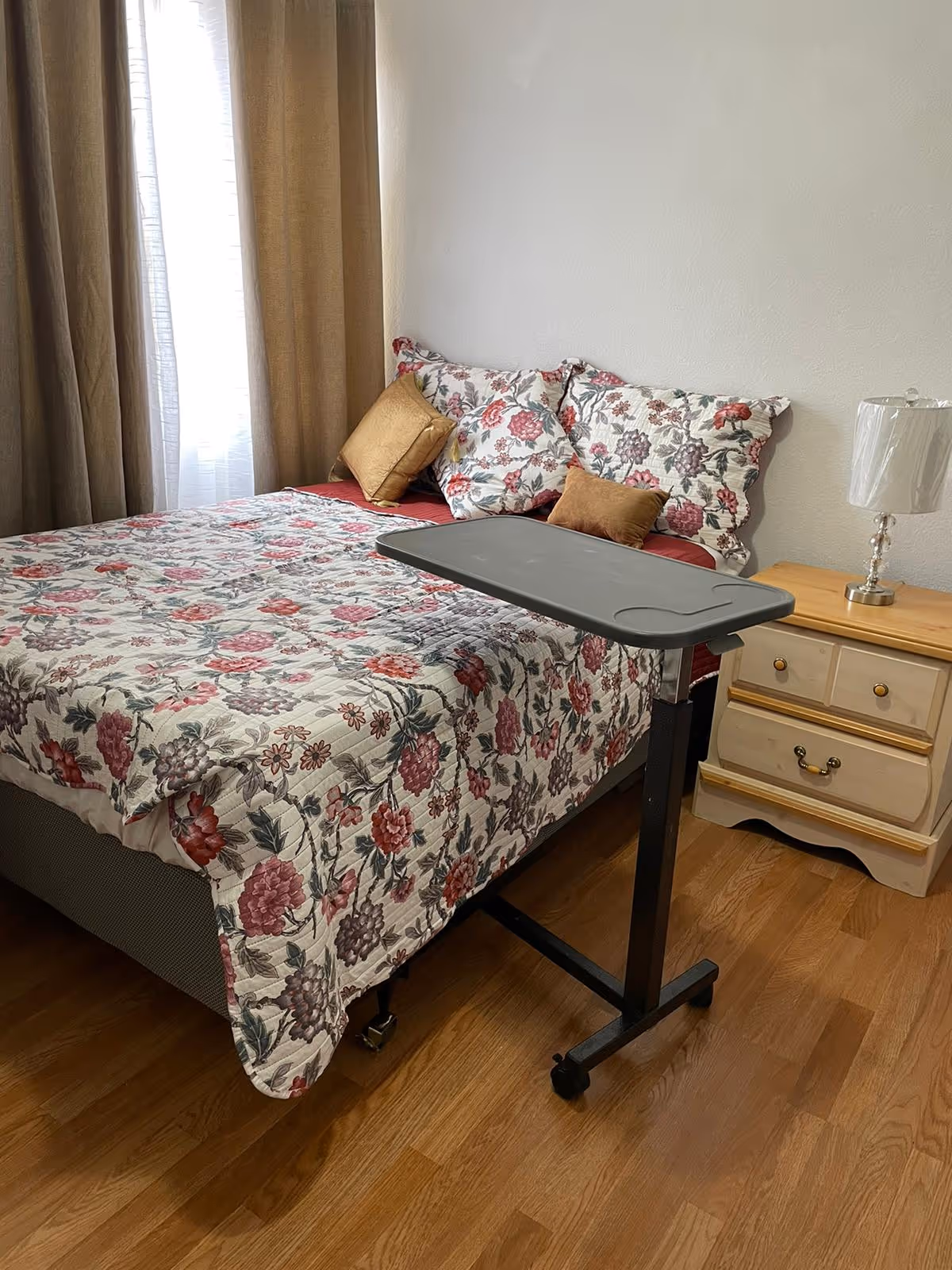 A bedroom with a bed covered in a floral patterned quilt and matching pillows, two golden accent pillows, a small overbed table on wheels, a wooden nightstand with two drawers, and a lamp with a white shade. Beige curtains cover a window in the background.