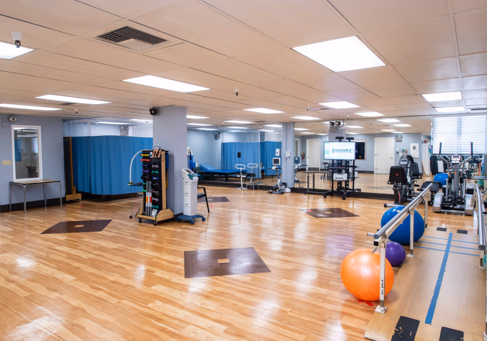 A spacious physical therapy or rehabilitation room with wooden floors, exercise balls, parallel bars, various exercise equipment, and blue privacy curtains. There is a large mirror on one wall and a TV screen displaying 'OmniVR II'.