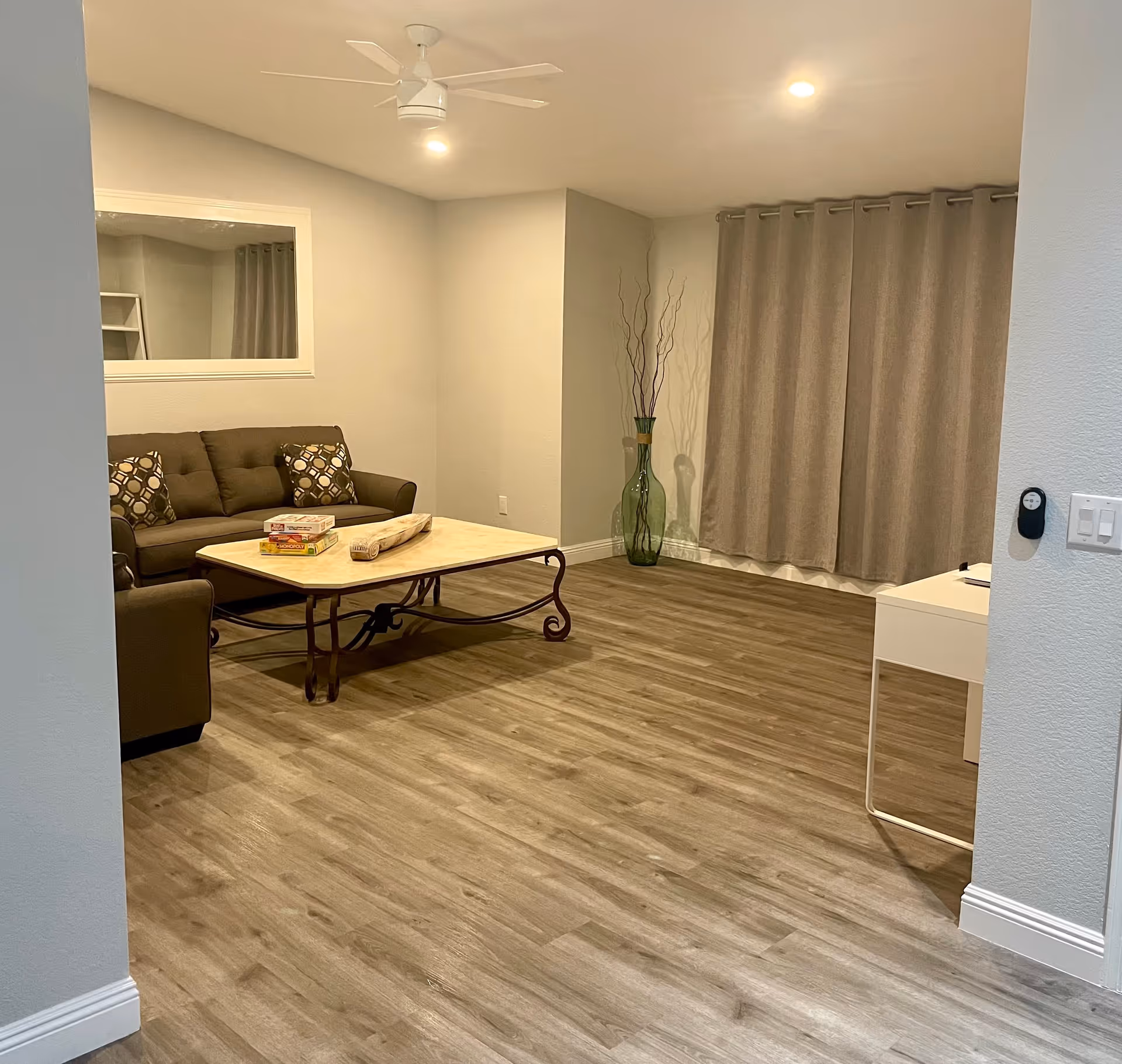 A cozy living room with a brown sofa adorned with patterned cushions, a large square coffee table with board games on top, a tall green vase with decorative branches in the corner, beige curtains covering a window, and a ceiling fan with lights. The room has wood flooring and neutral-colored walls.
