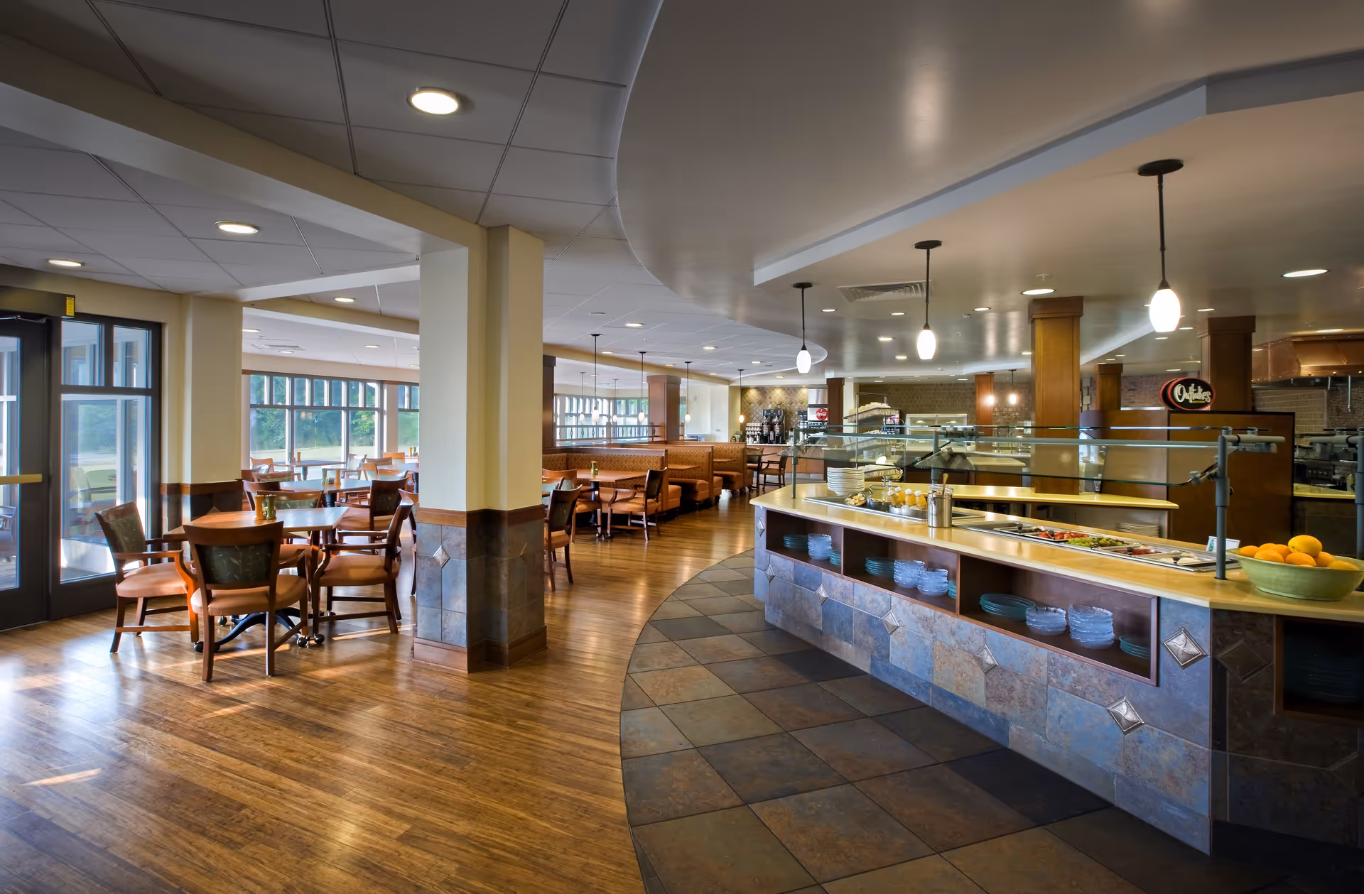 Interior view of a dining area in a senior living facility with wooden floors, multiple tables and chairs, a salad bar counter with plates and bowls, pendant lighting, and large windows letting in natural light.