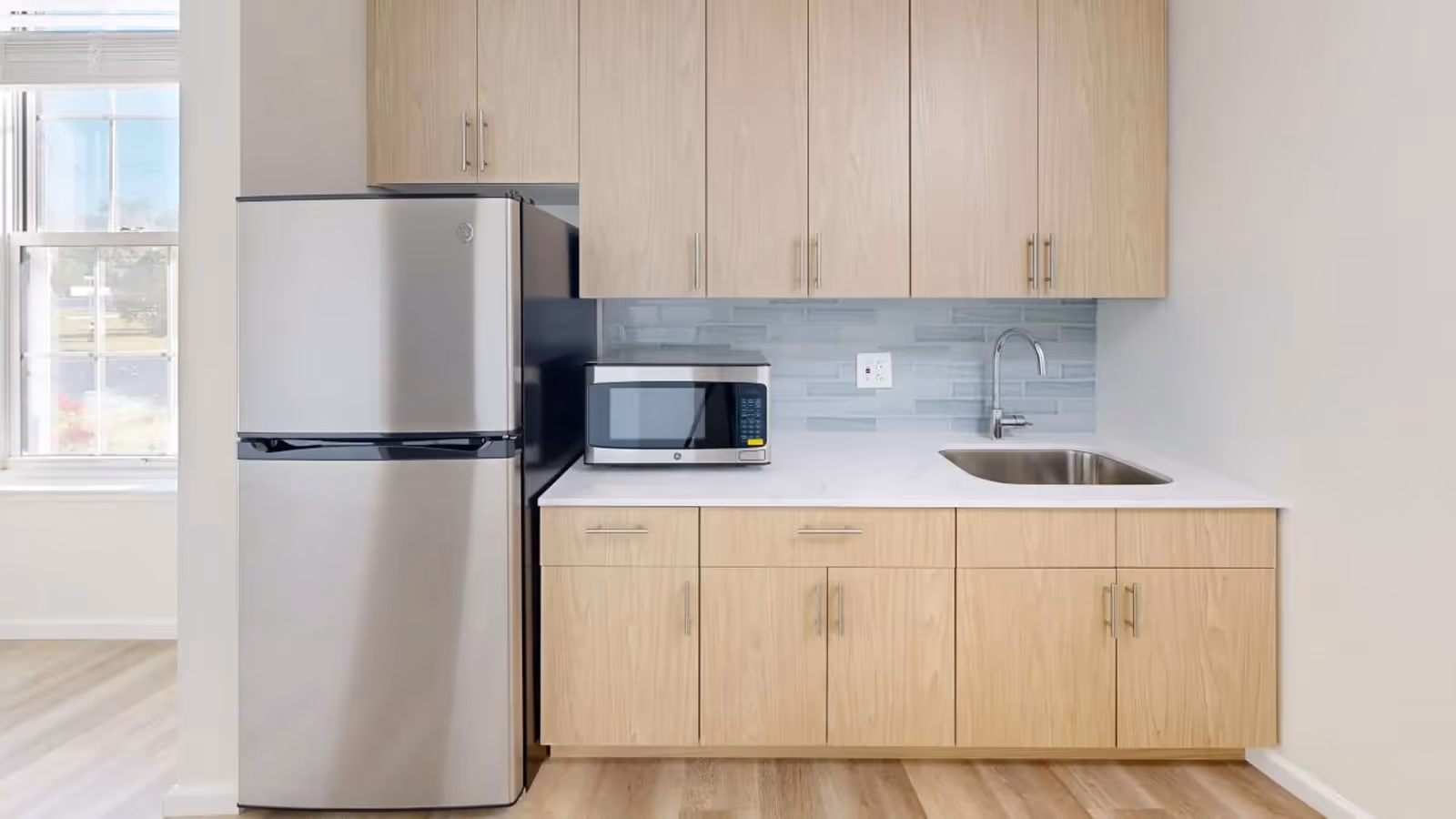 A modern kitchen area featuring light wood cabinets, a stainless steel refrigerator, a microwave on the countertop, a stainless steel sink with a faucet, and a light blue tiled backsplash. A window with blinds is visible to the left, allowing natural light into the space.