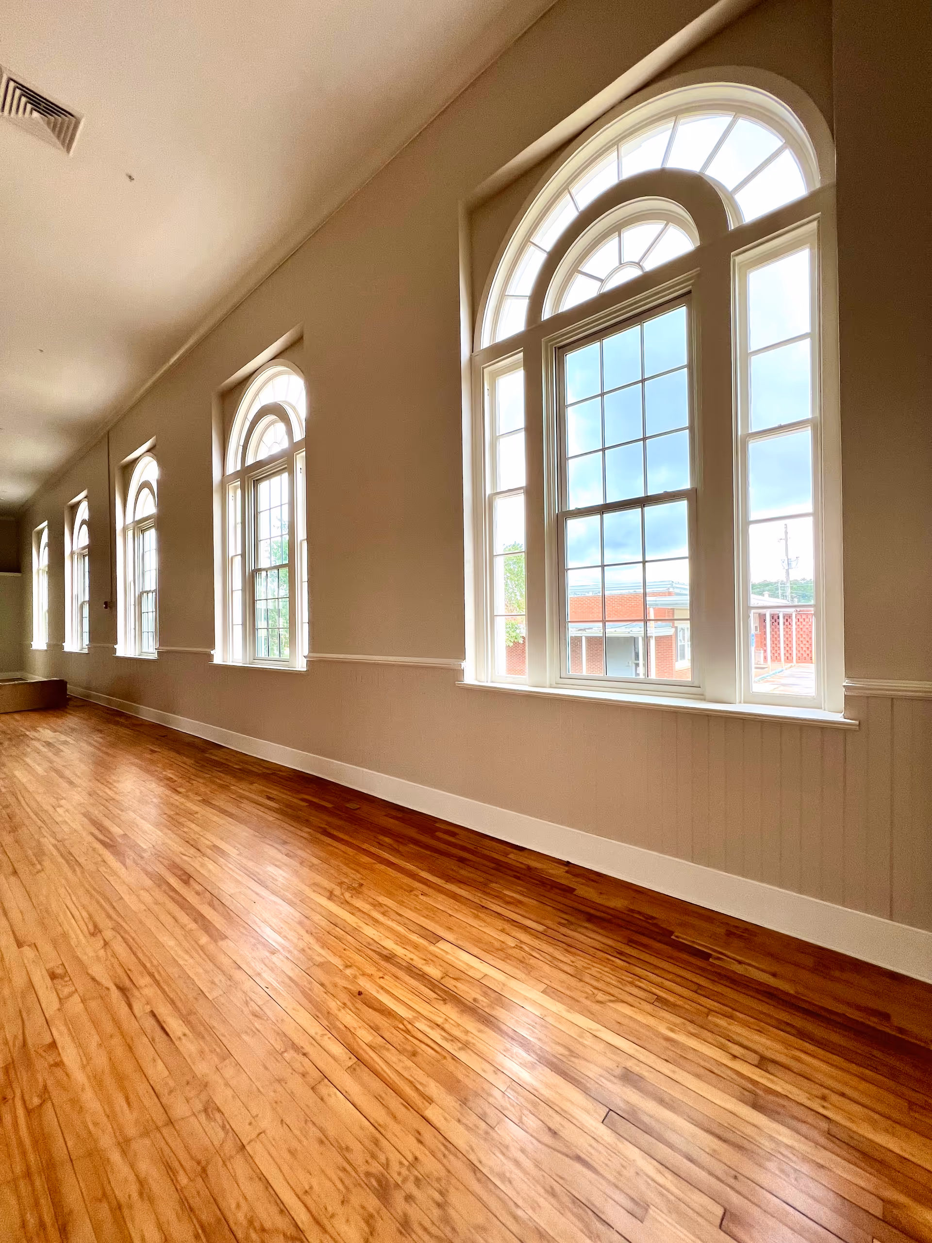 Sunlit interior hallway with polished hardwood floors and a row of tall arched windows along one wall.