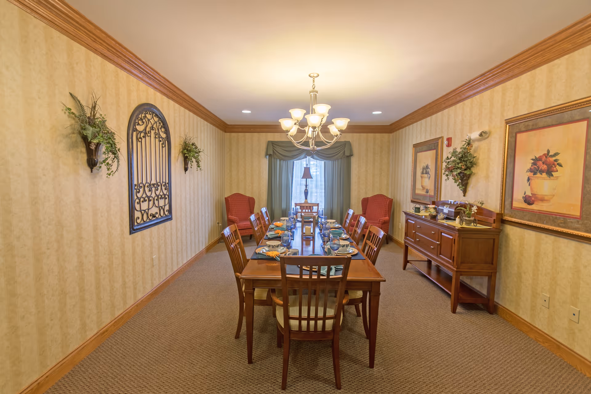 A formal dining room with a long wooden table set for eight people with plates, glasses, and napkins. The room has beige wallpaper with a subtle pattern, crown molding, and carpeted floor. Two red upholstered armchairs are placed near a window with green curtains at the far end. The walls are decorated with framed artwork, wall-mounted plants, and a decorative wrought iron piece. A wooden sideboard with additional decor is on the right side of the room, and a chandelier with multiple lights hangs above the table.