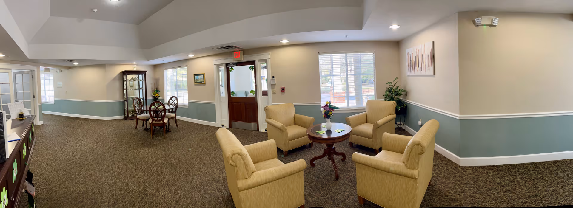 Spacious senior living facility lounge with upholstered armchairs around a central table, a small dining set, and an entry door and windows.