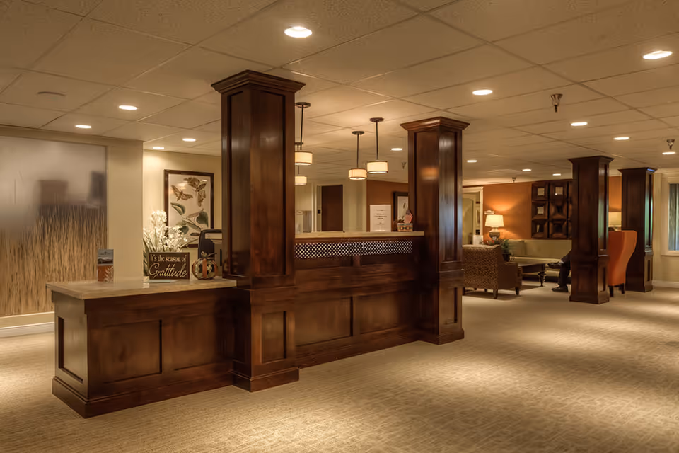 A warm and inviting reception area with a large wooden front desk featuring decorative columns. The space is softly lit with ceiling lights and pendant lamps. In the background, there is a seating area with armchairs and a sofa, along with framed artwork and a table lamp, creating a cozy atmosphere.