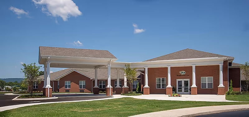 Front exterior of a red-brick nursing and rehabilitation center with columns and a covered drive-up entrance under a clear blue sky.