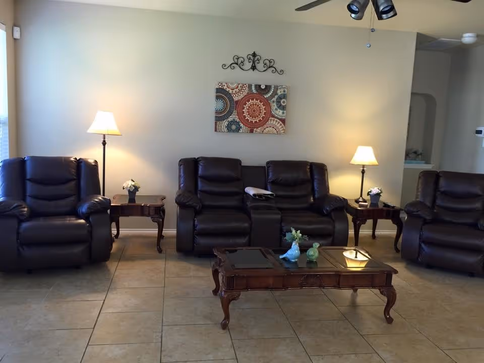 A living room with three dark brown leather recliners and a matching loveseat arranged around a wooden coffee table with decorative items. Two side tables with lamps and small flower pots flank the loveseat. A ceiling fan with lights is visible above, and a decorative wall hanging is centered on the beige wall behind the seating.