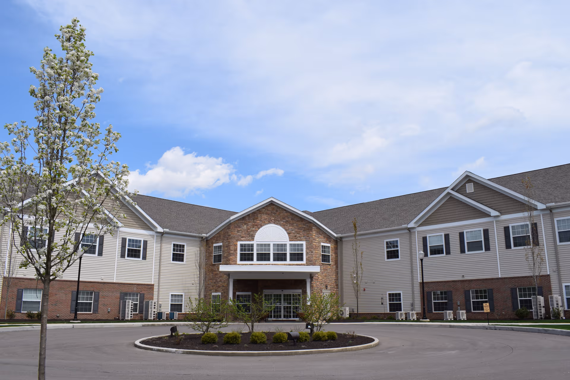 Front exterior view of a two-story senior living facility building with a circular driveway, landscaped bushes, and a tree with white blossoms under a partly cloudy blue sky.