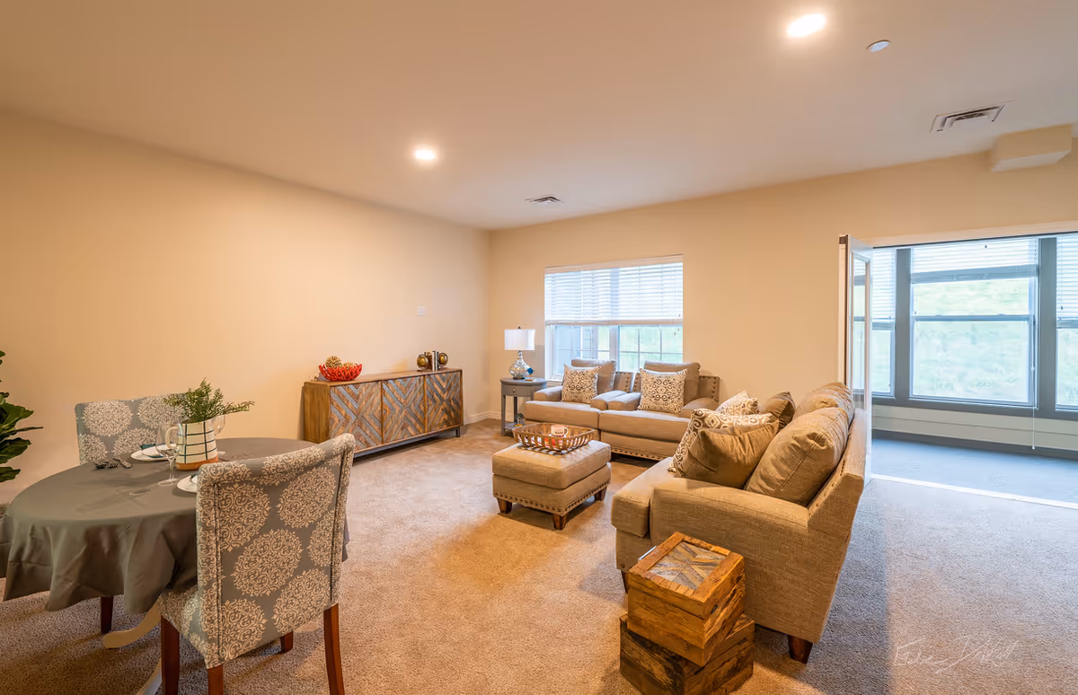 A cozy living room area with beige walls and carpet. The room features two beige sofas with patterned throw pillows, a matching ottoman, a wooden side table with a lamp, and a wooden cabinet against the wall. There is a small round dining table with two patterned chairs and place settings. Large windows with blinds allow natural light to enter, and a doorway leads to a sunroom or enclosed porch with additional windows.
