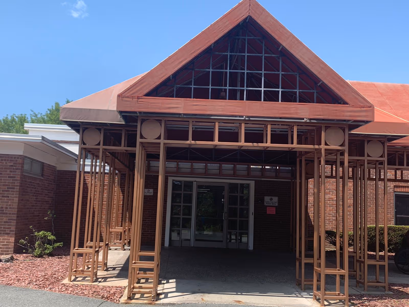Entrance of a brick building with a covered walkway supported by a metal frame structure painted brown. The roof of the walkway is triangular and covered with a reddish material. There are glass double doors at the entrance and some small bushes and landscaping with red mulch around the building.