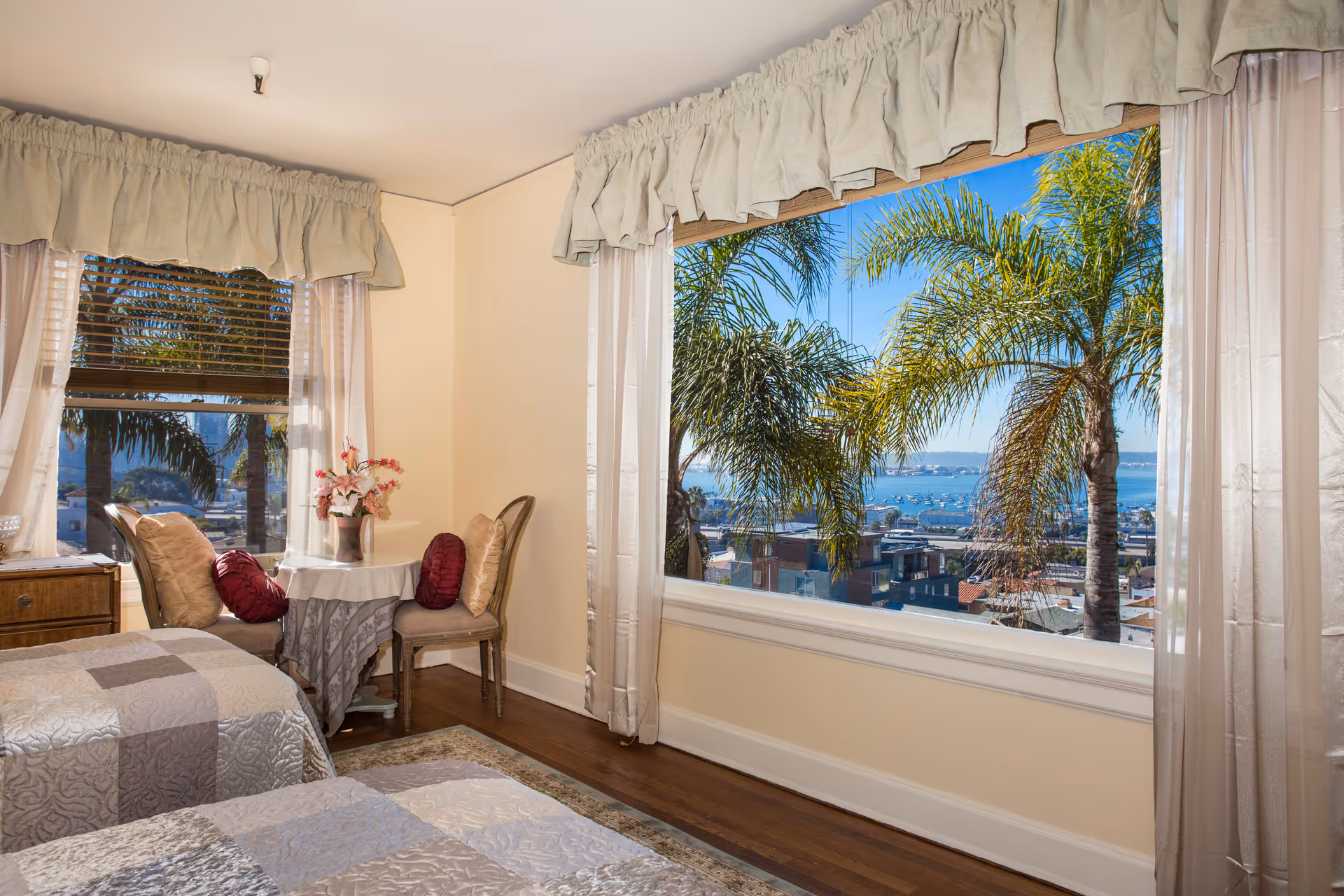 A bright bedroom with two twin beds covered in patterned bedspreads, a small round table with a vase of flowers, two chairs with decorative pillows, large windows with sheer curtains and valances, and a view of palm trees and a waterfront cityscape outside.