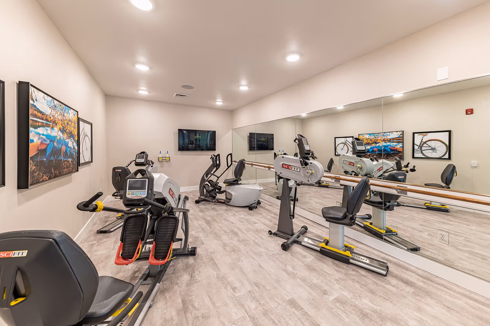 A small exercise room with several pieces of SciFit exercise equipment including recumbent bikes and elliptical machines. The room has light-colored walls, a large mirror covering one wall, framed pictures of bicycles and nature on the walls, and a television mounted on the far wall displaying the Cloverland Park Senior Living logo.