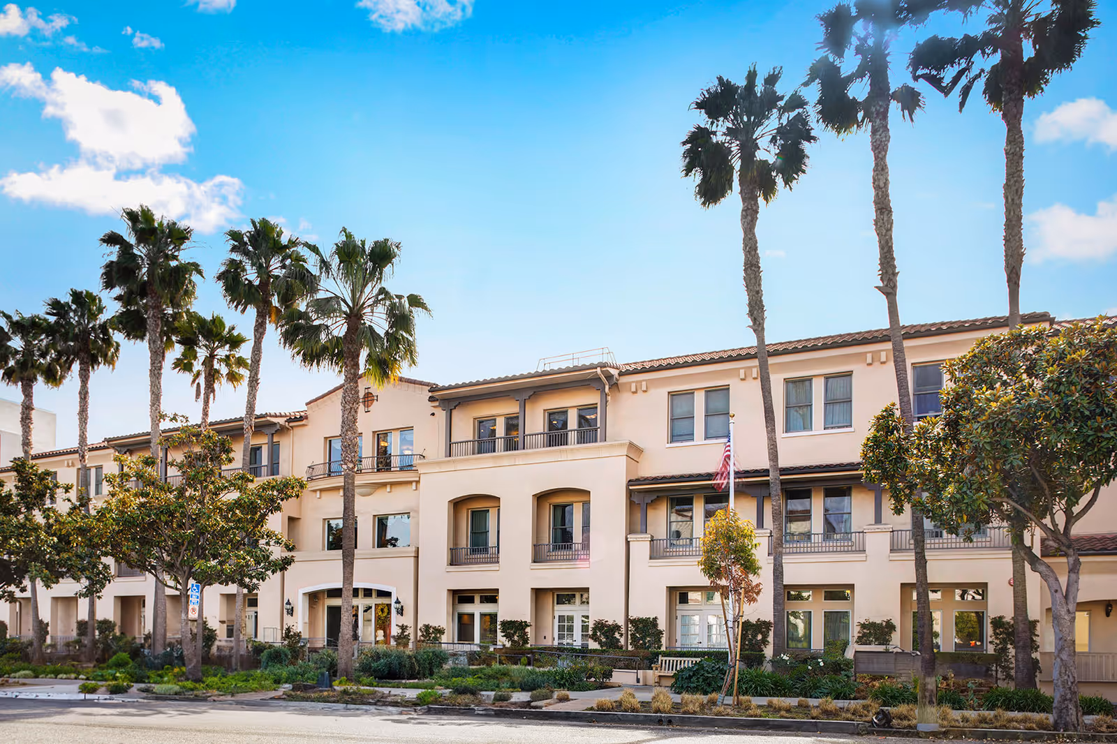 Exterior view of a three-story senior living facility building with beige walls and a tiled roof, surrounded by palm trees and other greenery under a blue sky with some clouds.
