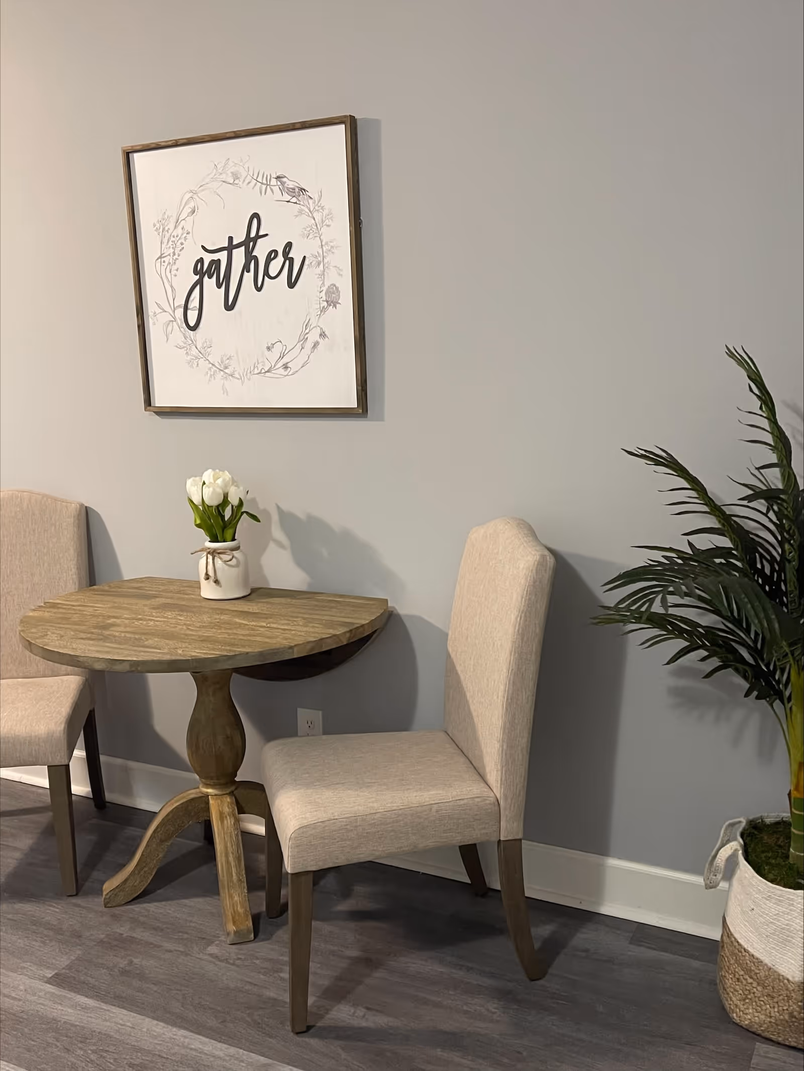 A small wooden round table with a vase of white tulips on top, flanked by two beige upholstered chairs. Above the table is a framed wall art with the word 'gather' surrounded by a floral wreath. To the right is a potted green plant in a woven basket. The setting is against a light gray wall with wood flooring.