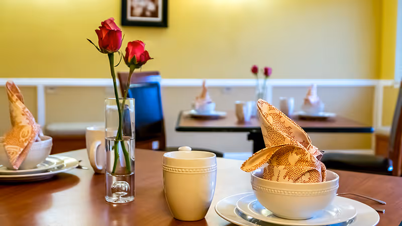 A dining table set with white plates, bowls, cups, and folded patterned napkins. A clear glass vase with two red roses is placed on the table. In the background, there are more tables similarly set with napkins and cups, against a yellow wall with white trim and a framed picture.