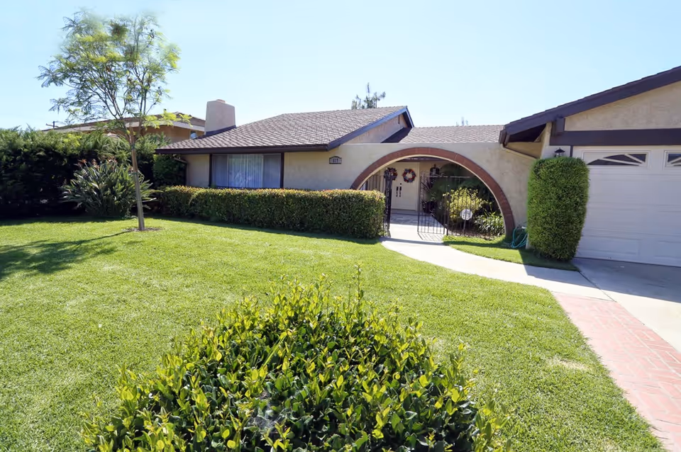 Front exterior view of a single-story house with a well-maintained green lawn, a small tree, bushes, and a driveway leading to a garage. The house features a distinctive arched entryway with a gate and two wreaths hanging on the front door.