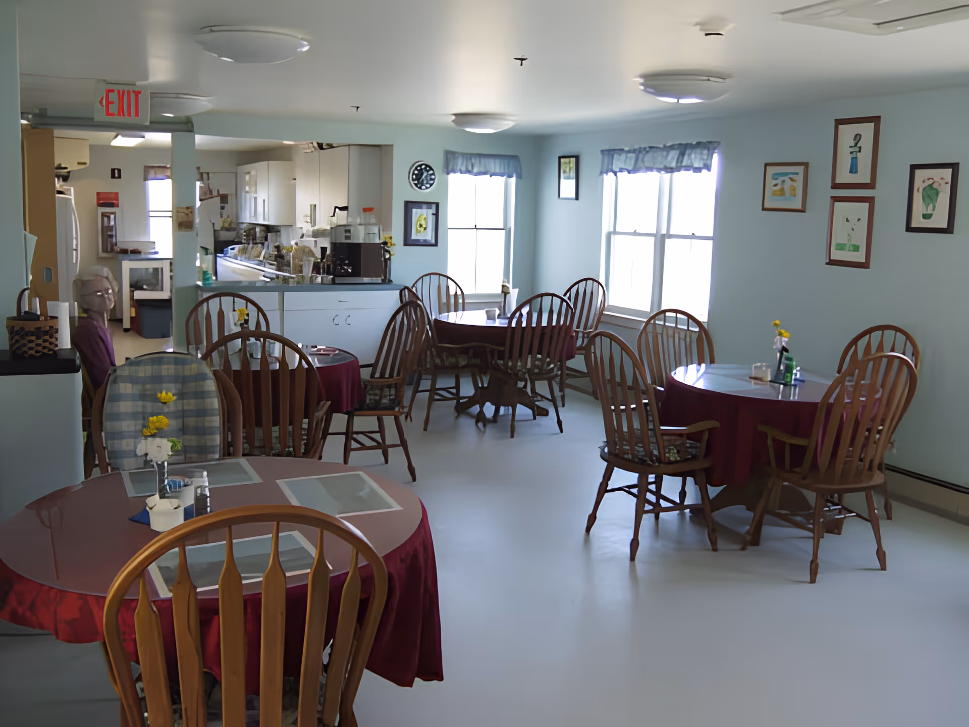 A dining room with several round tables covered with red tablecloths and wooden chairs around them. Each table has a small vase with yellow flowers and placemats. The room has light blue walls with framed pictures, two large windows with blue valances, and a kitchen area visible in the background. An elderly person is seated near the kitchen area. There is an exit sign on the ceiling near the kitchen.