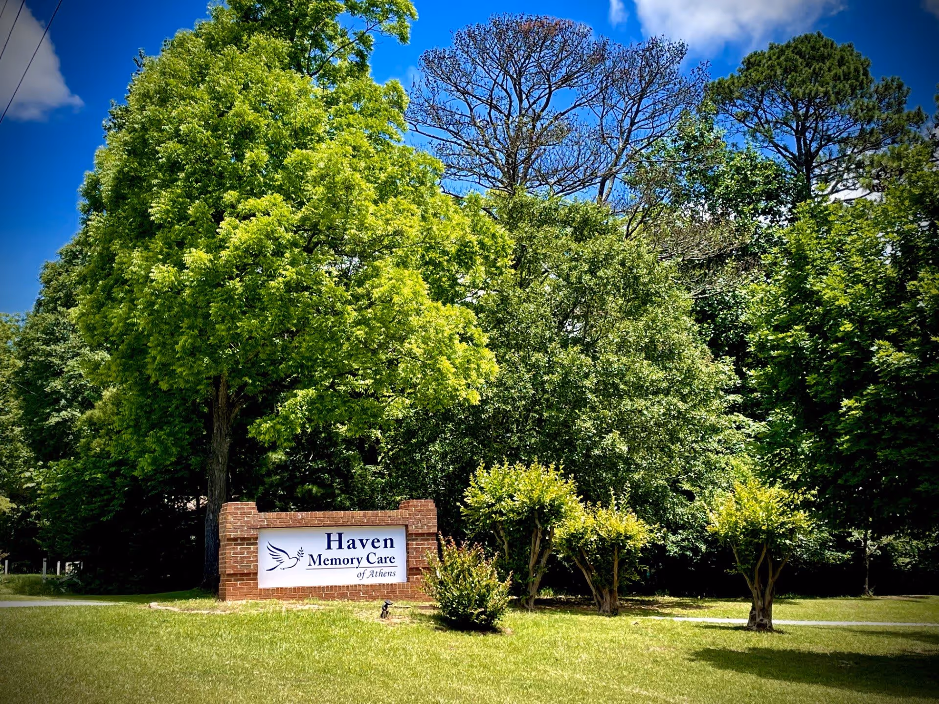 A brick sign for Haven Memory Care of Athens surrounded by green grass and various trees under a blue sky with some clouds.