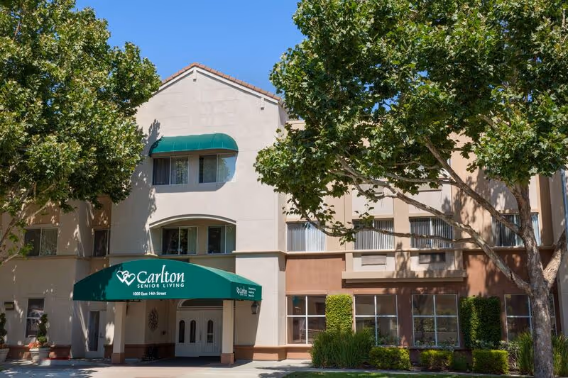 Exterior view of Carlton Senior Living San Leandro building with a green awning over the entrance, surrounded by trees and landscaping under a clear blue sky.