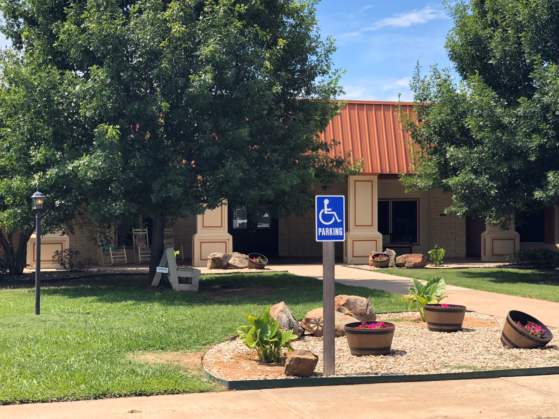 Exterior view of Mineral Wells Nursing & Rehabilitation facility showing a building with a red roof and beige walls partially obscured by green trees. In front, there is a landscaped area with rocks, plants, and flower pots, along with a blue handicapped parking sign on a post. A sidewalk leads to the entrance of the building.