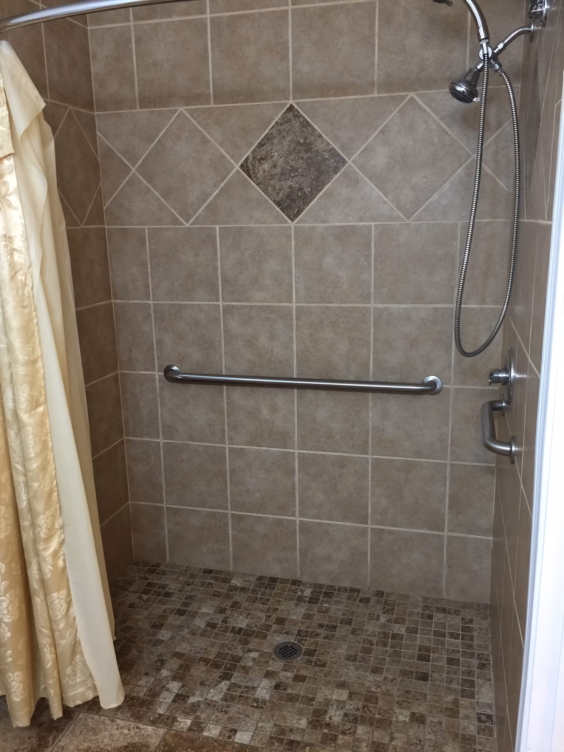 A tiled shower area with a beige and brown color scheme. The walls have large square tiles with a decorative diamond-shaped tile in the center. The floor has smaller mosaic tiles. There is a metal grab bar mounted horizontally on the wall and a handheld showerhead attached to a flexible hose. A beige shower curtain is partially visible on the left side.