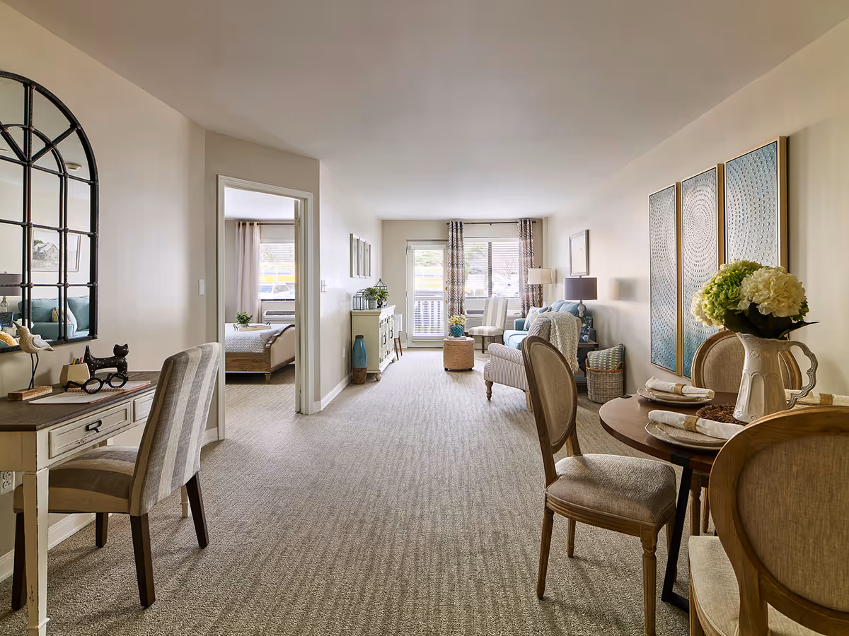 A bright and spacious senior living apartment interior featuring a dining area with a round table set for four, a cozy living room with armchairs and a sofa, and a bedroom visible through an open doorway. The room is decorated with neutral tones, patterned curtains, wall art, and a large mirror above a desk with a striped chair.
