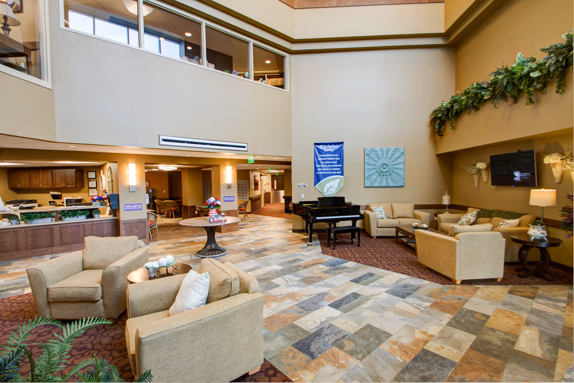 A spacious and well-lit senior living facility common area with comfortable beige armchairs and sofas arranged around coffee tables. There is a black grand piano against the far wall, decorative wall art, and a flat-screen TV mounted on the wall. The floor is tiled with a mix of earth tones, and there are plants and floral arrangements adding greenery to the space. The area also features a reception desk and an open hallway leading to other rooms.