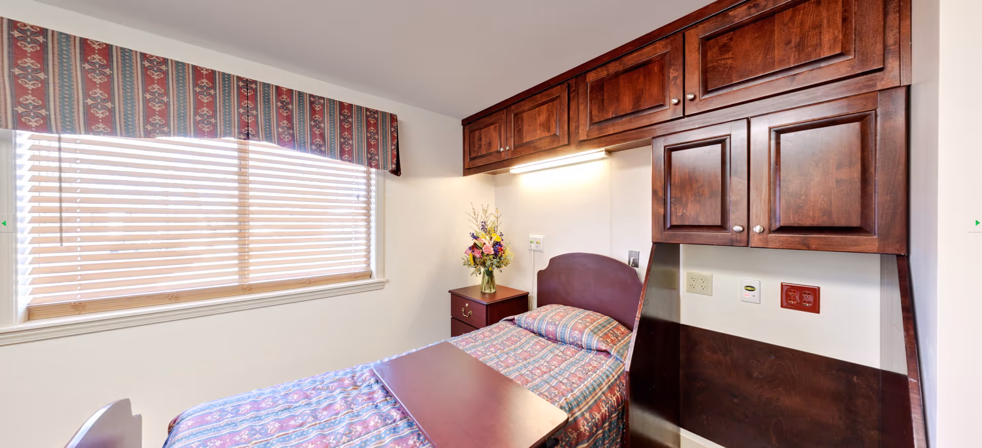 A small bedroom with a single bed covered in a patterned bedspread. Above the bed are wooden cabinets mounted on the wall. Next to the bed is a nightstand with a vase of colorful flowers. A window with blinds and a decorative valance lets in natural light.