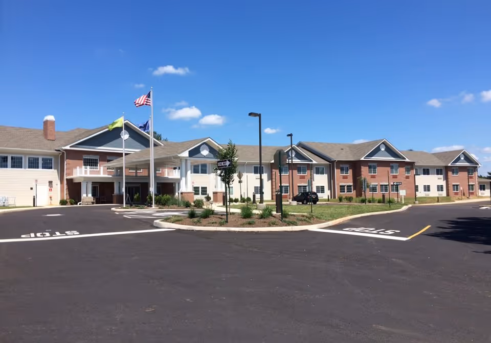 Exterior view of Morningside House of Collegeville, a two-story senior living facility with a combination of brick and light-colored siding. The building has multiple windows, a covered entrance with columns, and three flagpoles displaying flags. The foreground shows a paved driveway with stop markings and landscaped areas with small trees and shrubs under a clear blue sky.
