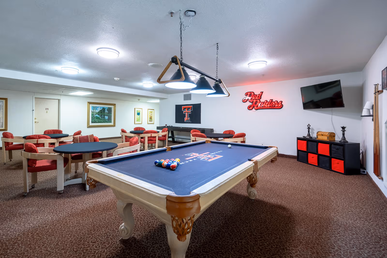 Indoor community game room with a pool table, round card tables and red chairs, a wall-mounted TV and a 'Red Raiders' neon sign.