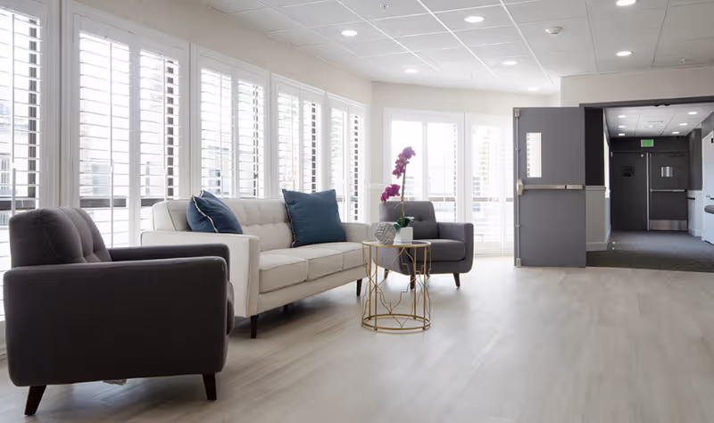 A bright and modern living room area with a beige sofa adorned with blue pillows, two gray armchairs, and a small round gold coffee table with a purple orchid and decorative items. Large windows with white shutters allow natural light to fill the space. A hallway with gray doors is visible in the background.