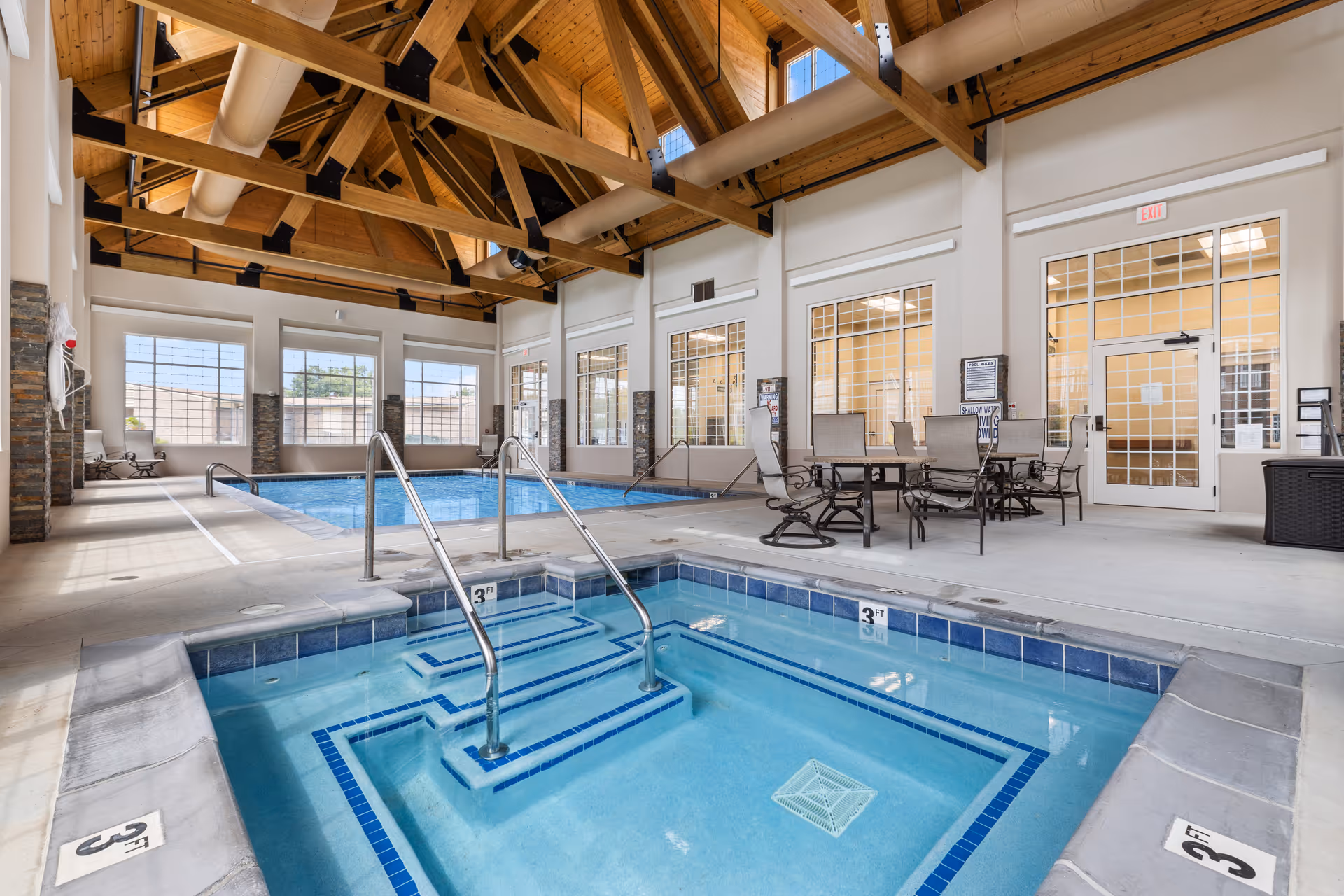Indoor pool area with a hot tub in the foreground and a larger swimming pool in the background. The space features high wooden ceilings with exposed beams and large windows letting in natural light. There are tables and chairs for seating along the side near glass doors and windows.