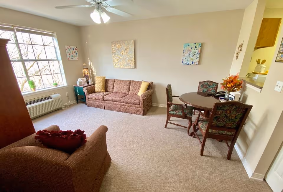 A cozy senior living room with a large window letting in natural light. The room features a brown patterned sofa with two yellow cushions, a matching armchair with a red pillow, a round wooden table with four floral upholstered chairs, and a small side table with decorative items. The walls are decorated with three colorful paintings, and there is a ceiling fan with lights overhead.