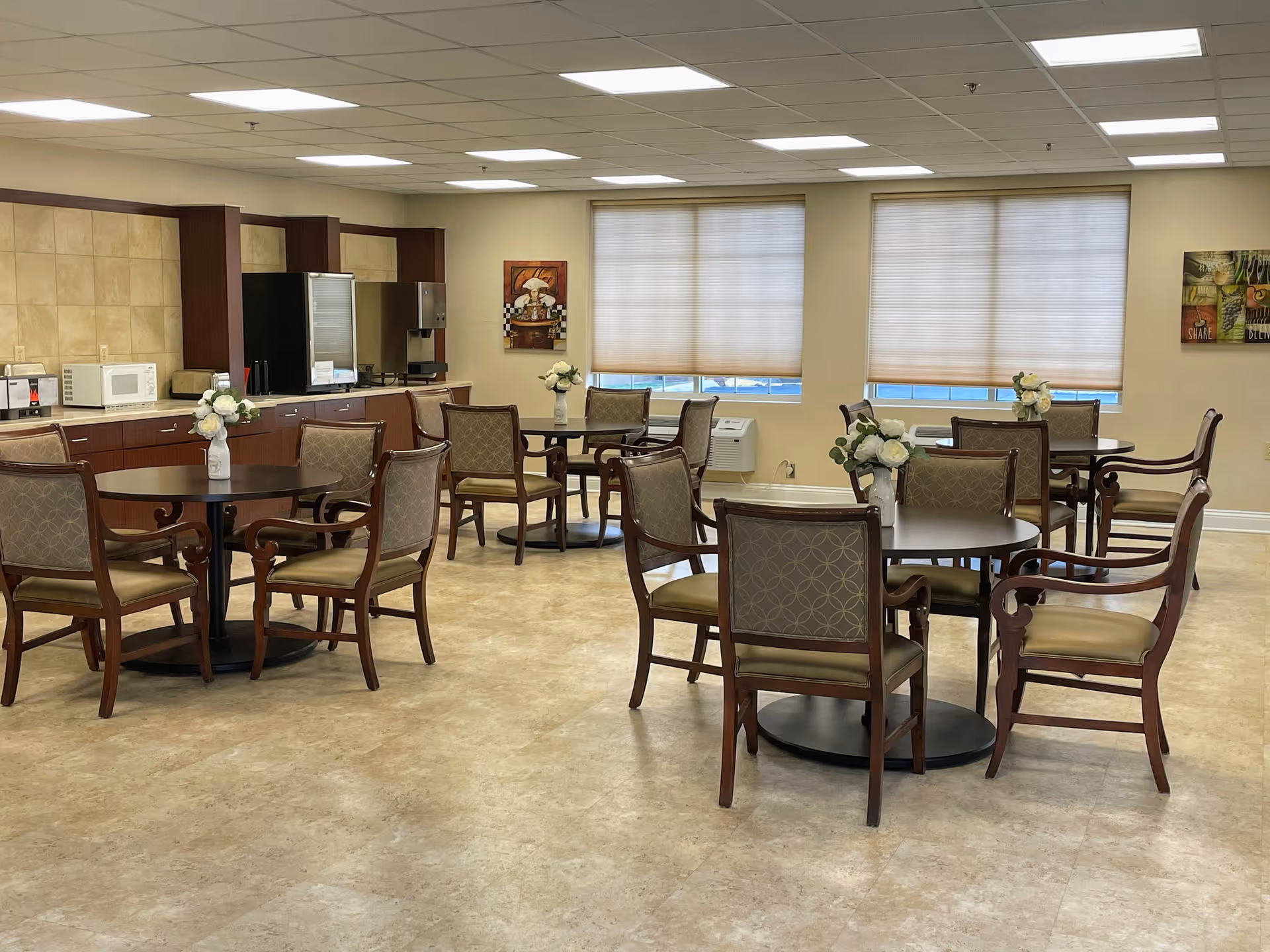 A spacious dining area with several round tables, each surrounded by four wooden chairs with cushioned seats and backs. Each table has a small vase with white flowers. The room has large windows with blinds partially drawn, beige walls, and a tiled floor. Along one wall, there is a kitchenette area with a microwave, coffee machine, and other appliances. The ceiling has recessed lighting panels.